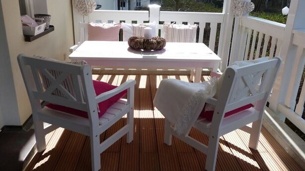 Um deck de madeira com mesa e cadeiras brancas, guarda-corpo branco e vista para edifícios ao fundo.
