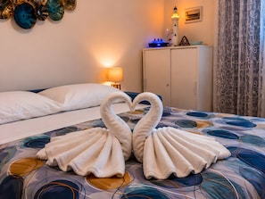A bed with a decorative towel arrangement in the shape of two swans, a nightstand with a lamp, and a wall-mounted clock. - M.a.g.a. Holiday House in Taormina – Studio with Parking, Sea View, and Self Check-in (Taormina)