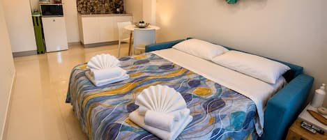 A hotel room with a bed, a small table, a chair, and a kitchenette in the background.