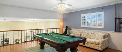 A pool table in a room with a ceiling fan, a sofa, and a window with blinds.