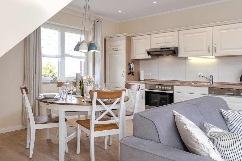 Une cuisine moderne avec des meubles blancs, une table à manger accompagnée de chaises, et un canapé gris dans l’espace salon.