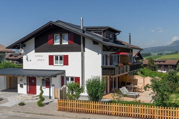 A two-story building with a balcony, a wooden fence, and a clear view of the surrounding landscape. - Holiday home with 1 bedroom (Obermaiselstein)