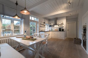Une cuisine avec des meubles blancs, une table à manger avec des chaises, des luminaires suspendus et une vue sur l’extérieur grâce à de grandes fenêtres.