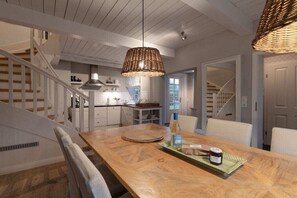 Ein Essbereich mit einem Holztisch, Stühlen, einer gewebten Pendelleuchte und Blick auf eine Treppe, die zur Küche führt.