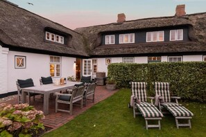Ein traditionelles Haus mit einem Reetdach, einer Terrasse, Sitzgelegenheiten im Freien und einem Hecken.