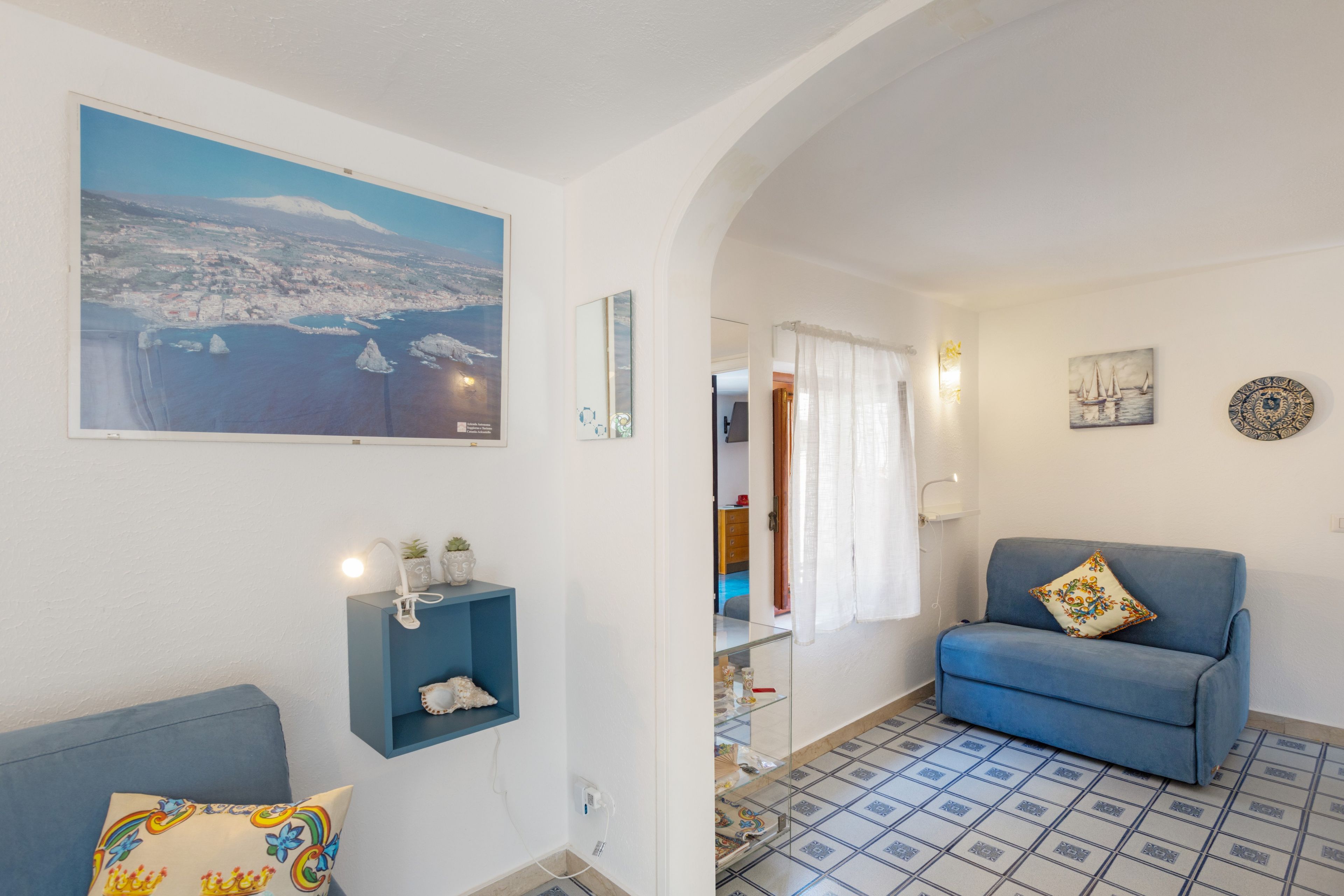 Un salon avec un canapé, un mur d’accent bleu, un tableau représentant un paysage côtier et un sol carrelé.