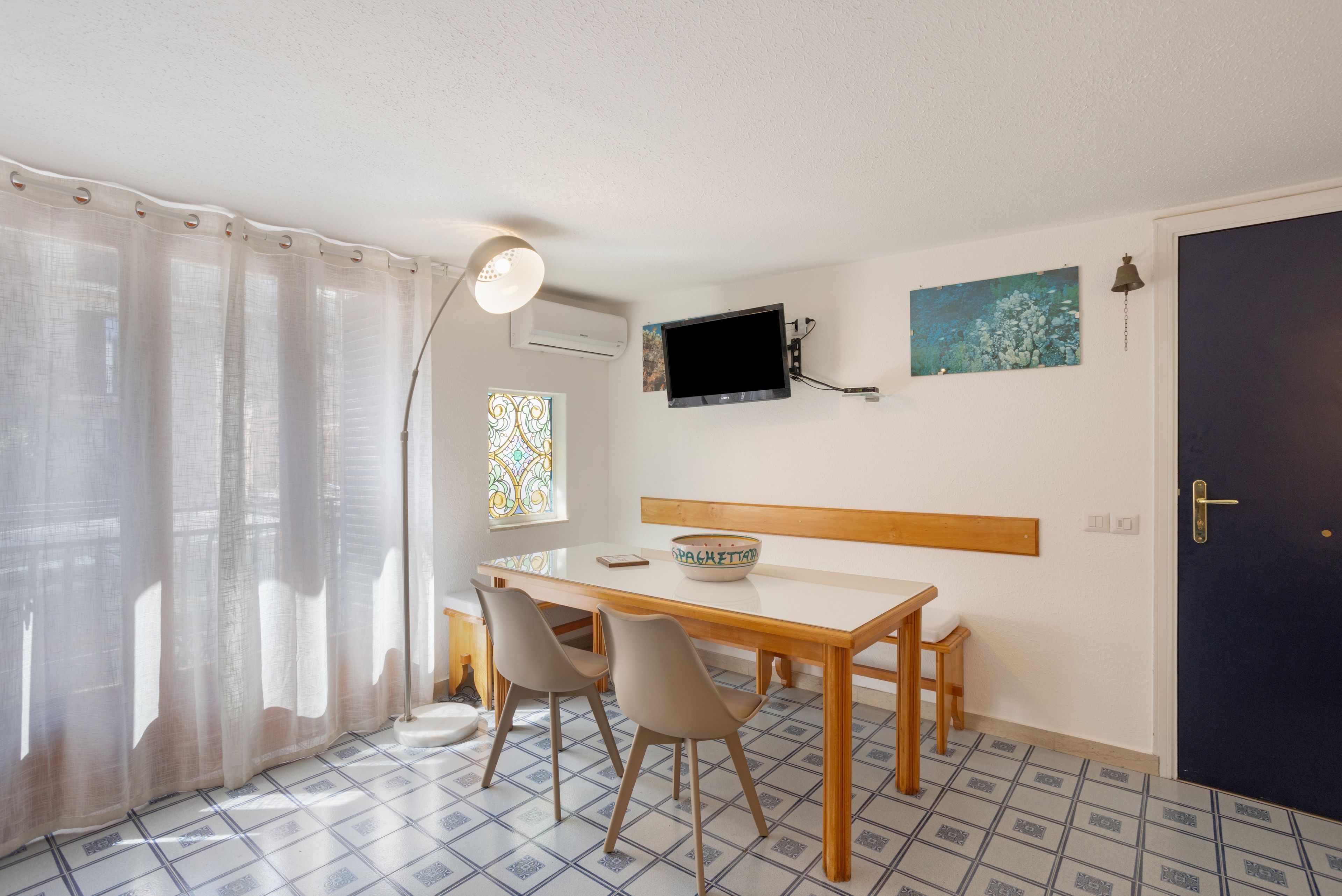 Una habitación con una mesa de comedor, sillas, un televisor montado en la pared, una ventana con cortinas y una puerta.