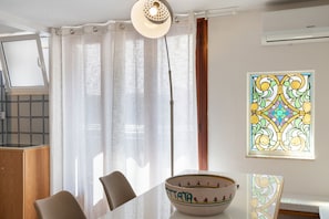 A room with a dining table, chairs, a window with curtains, a wall-mounted air conditioner, and a stained glass window. - Agatella's House Apartment with Sea View, Private Terrace, and Air Conditioning (Aci Castello)