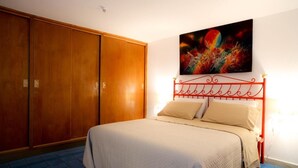 Quarto de hotel com cama, cabeceira de metal vermelha, guarda-roupa de madeira grande e um quadro abstrato colorido na parede.