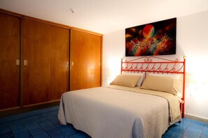 Une chambre d’hôtel avec un lit, une tête de lit en métal rouge, une grande armoire en bois et un tableau abstrait aux couleurs vives accroché au mur.
