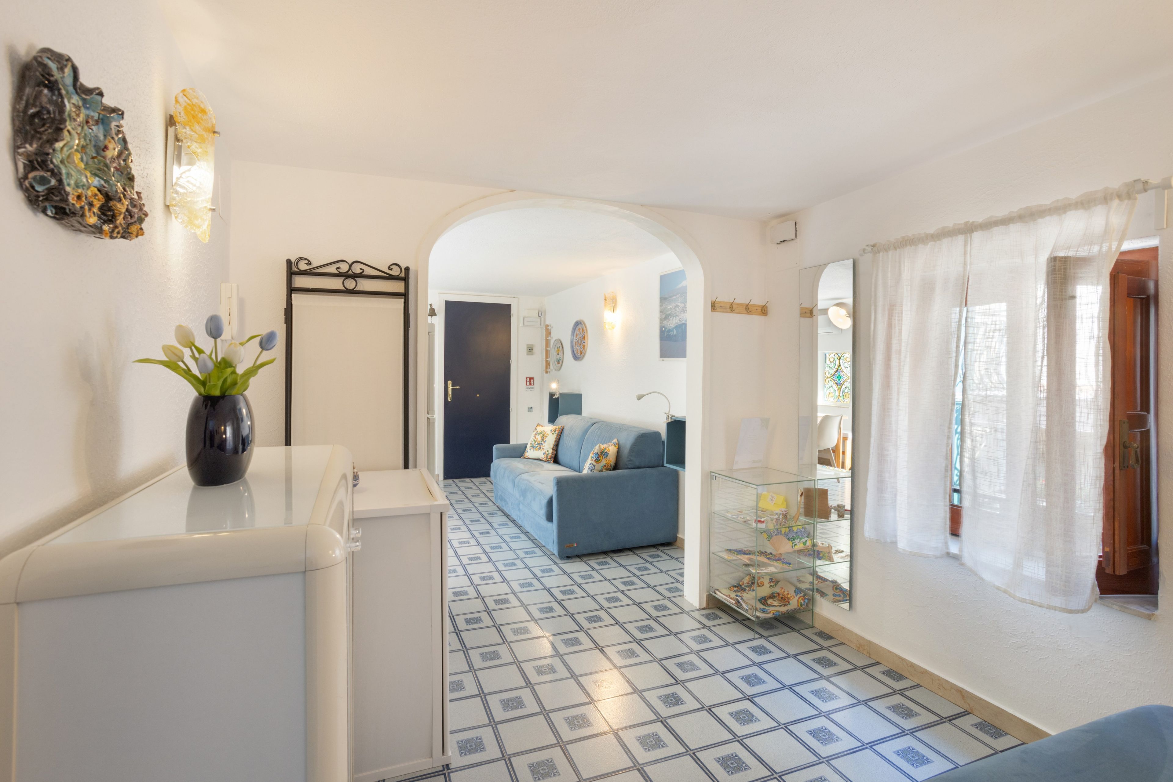 A hallway with a tiled floor, a sofa, a glass display case, and a door leading to a room.