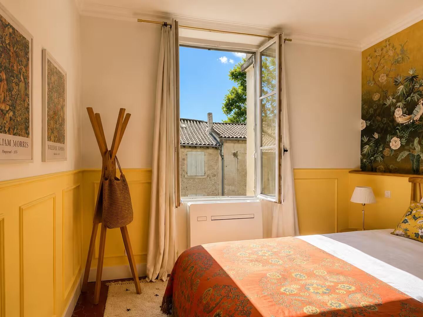 A hotel room with a bed, a window with curtains, a wall with a painting, and a wooden rack.
