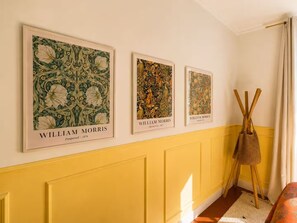 A hallway with three framed botanical prints on the wall, a yellow wall, and a woven basket on a stand. - Le Vernet – Avignon City Center – Air-Conditioned (Avignon)