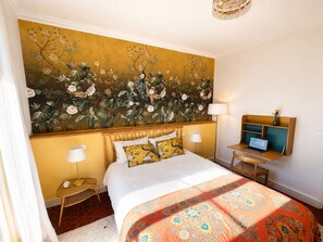 A hotel room with a large floral mural, a bed with a patterned bedspread, bedside lamps, a desk with a laptop, and a small table with a vase. - Le Vernet – Avignon City Center – Air-Conditioned (Avignon)