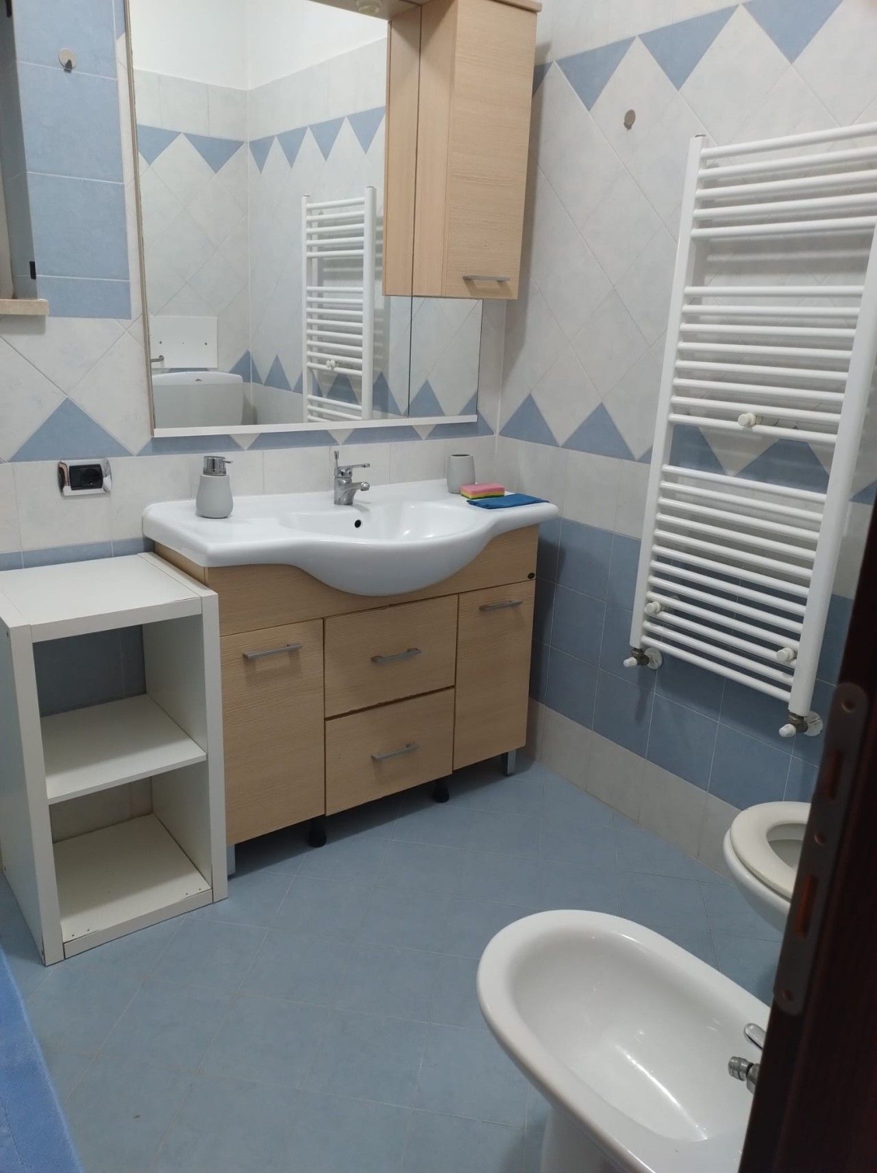 Casa de banho com lavatório branco, espelho e banheira branca. Inclui um porta-toalhas e um móvel com gavetas.