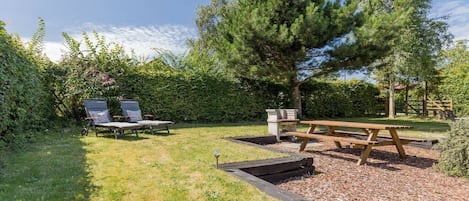 A garden with a wooden picnic table, two lounge chairs, and a large tree.