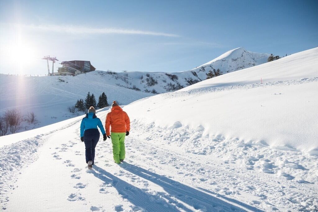Un sentier de montagne enneigé, parsemé d’empreintes, un ciel dégagé et un bâtiment visible au loin.