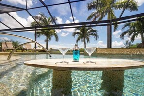 Ein Poolbereich mit Tisch und zwei Martini-Gläsern, umgeben von Palmen und blauem Himmel.