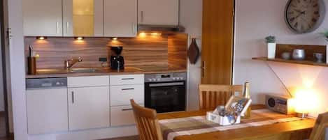 A compact kitchen with modern appliances, a dining table with chairs, and a wall clock.