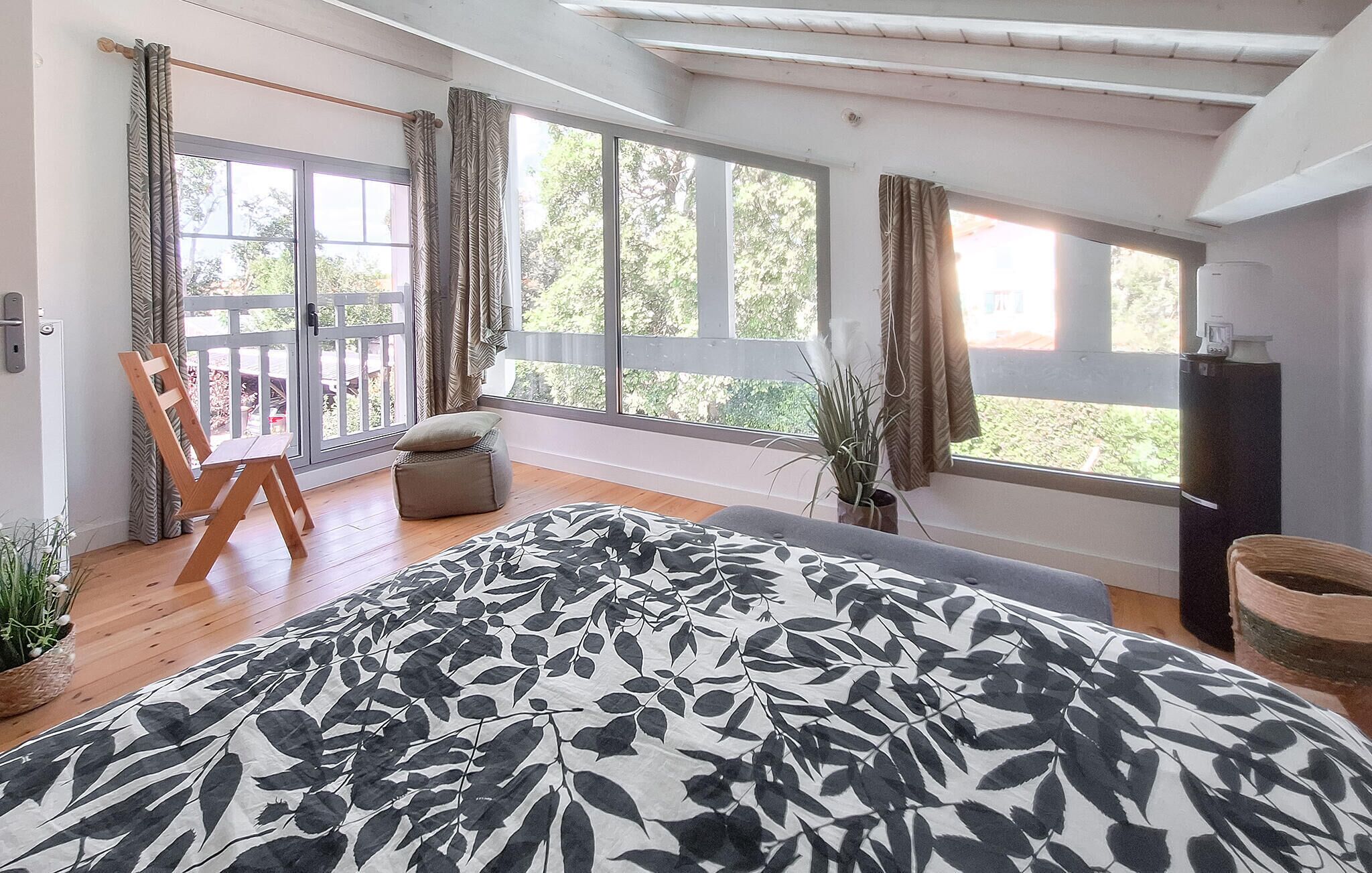 Une chambre à coucher avec un lit, une chaise, un petit tabouret, une plante en pot et une grande fenêtre avec des rideaux.
