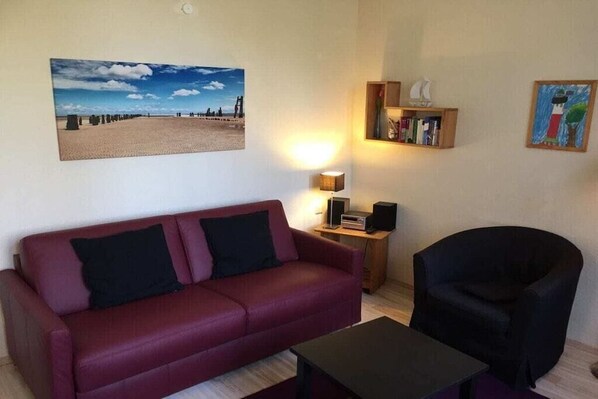 A living room with a red sofa, a black armchair, a coffee table, a bookshelf, and a painting on the wall. - Sandrose Villa am Park (Wangerooge)
