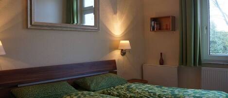 Une chambre avec deux lits, une table de chevet avec une lampe, un miroir au mur et une fenêtre avec des rideaux.