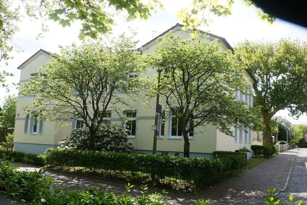 A two-story building with a light-colored facade, surrounded by greenery and a paved pathway.
