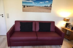 Ein rotes Sofa mit schwarzen Kissen, eine Lampe auf einem Holztisch und ein Gemälde mit Strandmotiv an der Wand.