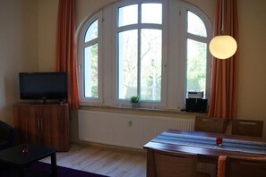 A room with a large arched window, a television on a wooden cabinet, a dining table with chairs, and a hanging light fixture. - Sandrose Villa am Park (Wangerooge)