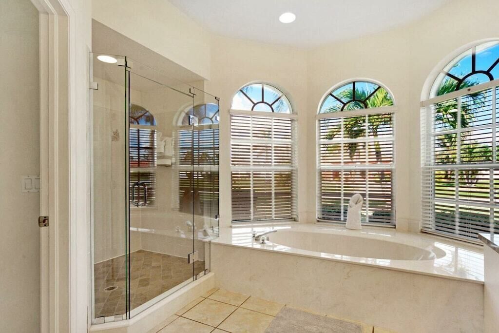 Une salle de bain dotée d’une douche à parois de verre, d’une grande baignoire et de trois fenêtres en arc de cercle équipées de stores.