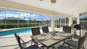 Um pátio externo com uma mesa de jantar posta para quatro pessoas, uma churrasqueira e uma piscina ao fundo.