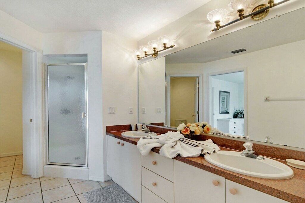 Un bany amb un envà de banyera de vidre, un lavabo doble i un mirall gran amb llums a sobre.