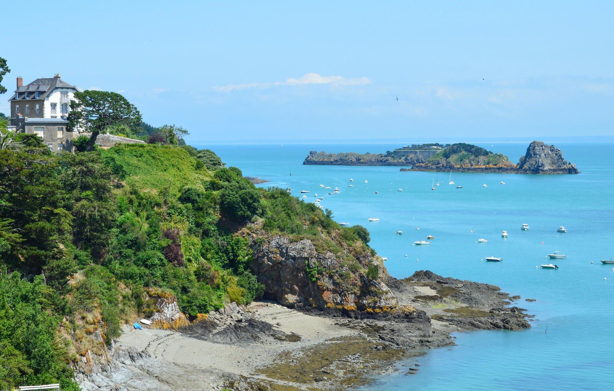 Paisagem costeira com uma casa numa colina com vista para o mar, costa rochosa e barcos na água.