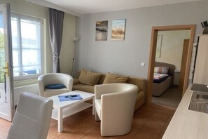 A modern living room with a sofa, armchairs, and a coffee table. There is a sliding door leading to a bedroom with a bed and a bathroom. - holiday home at the harbor (Zingst)