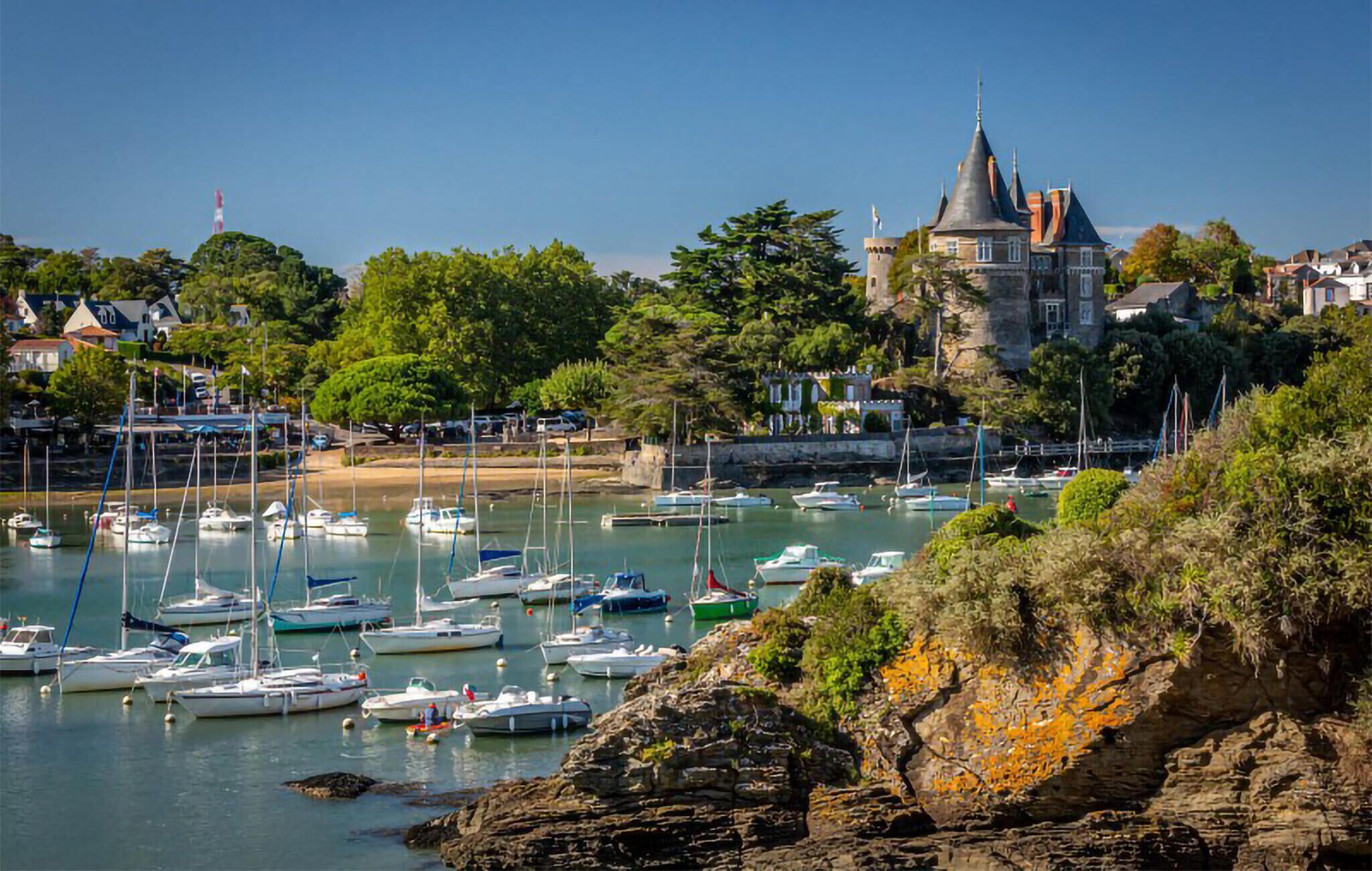 Un port où sont amarrés de nombreux bateaux, un imposant bâtiment qui ressemble à un château, et une végétation luxuriante tout autour.