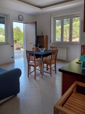 Ein Zimmer mit Esstisch, Stühlen, einer Wanduhr, einem Fenster mit Blick auf Grünflächen und einer Heizung.