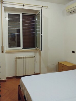 Ein Schlafzimmer mit einem Bett, einem Heizkörper, einem Fenster mit Jalousien und einem Holzschrank.