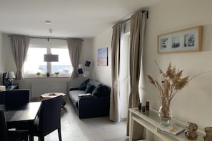 Ein modernes Wohnzimmer mit einer Couch, einem Esstisch mit Stühlen, einer Lampe und einer Vase mit Trockenpflanzen.