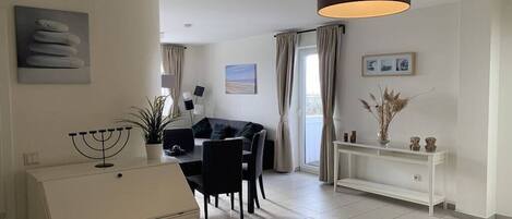 A modern living room with a dining area, a white console table, a black sofa, and a large window with curtains.