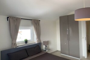 Ein Schlafzimmer mit einem Bett, einem Nachttisch mit Lampe, einem Kleiderschrank und einem Fenster mit Vorhängen.