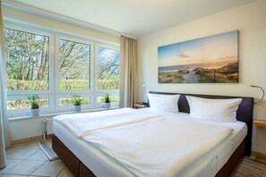 A hotel room with a large bed, a window with a view of trees, and a painting on the wall. - Anchorage - Binz Stars (Ostseebad Binz)
