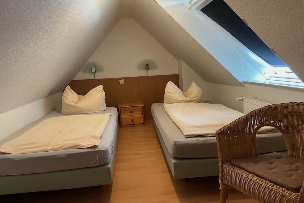 Een klein, driehoekig slaapkamer op zolder met twee bedden, een houten nachtkastje, een rieten stoel en een dakraam.