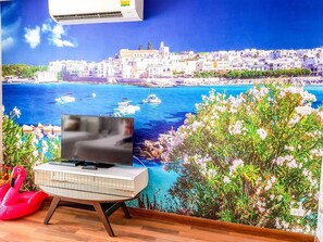 A room with a television on a stand, a red inflatable chair, and a wall mural of a coastal cityscape. - E202 Cozy 1-bedroom apartment with Playground near City (Nongkae)