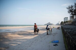 Ein Strand mit einem Pferd und Reiter, eine Person, die auf einem Pferd sitzt, und einem blauen Boot im Sand.