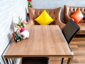Een houten tafel met een zwarte stoel, een bruine leren bank met gele en oranje kussens, en een witte bakstenen muur.