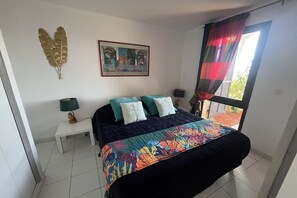 Ein Schlafzimmer mit einem Bett, einem Nachttisch, einer Lampe, einem Fenster mit Vorhängen und einem Wandbild.