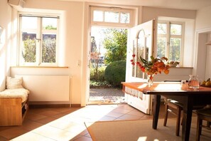Een kamer met een groot raam, een houten bank, een eettafel met stoelen en een vaas met bloemen.