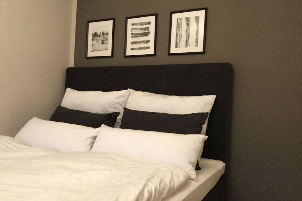 A neatly made bed with white and black bedding, a dark headboard, and three framed artworks on the wall above.