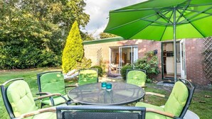 En udendørs patio med et rundt bord og fire stole, en grøn parasol og en murstensbygning i baggrunden.
