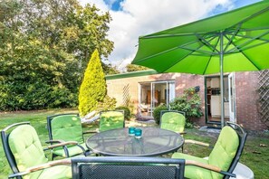 Eine Außenpatio mit rundem Tisch und vier Stühlen, einem grünen Sonnenschirm und einem Backsteinhaus im Hintergrund.